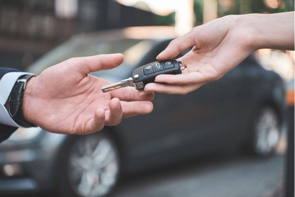 Closeup of hands passing off car keys, with a blurred background of a car.